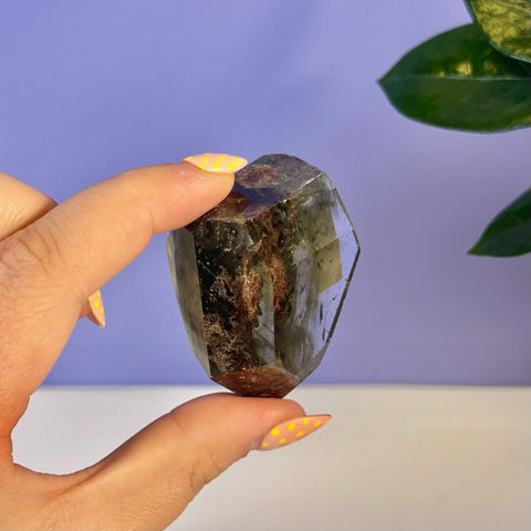 Hand holding a clear garden quartz free form with dark green and reddish-brown inclusions.