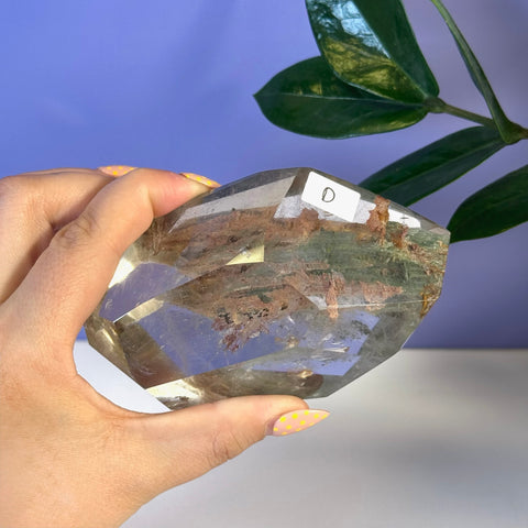 Faceted Garden Quartz free form crystal, clear with green and reddish-brown inclusions, held in hand.