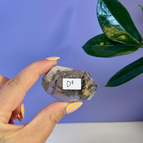 Hand holding a clear garden quartz free form crystal with dark inclusions and a D3 label.