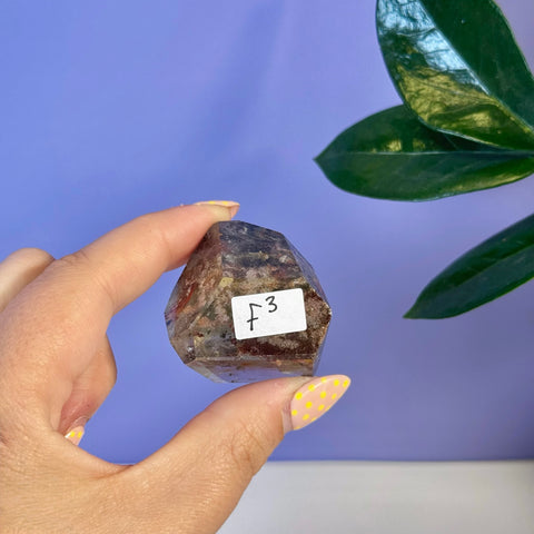 Garden Quartz Free Form F3, a clear crystal with earthy red and green inclusions, held in hand.