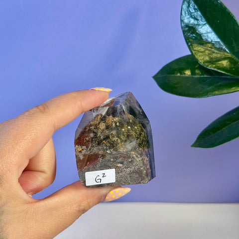 Clear Garden Quartz free form crystal with green and brown inclusions, held in hand. Variant G2.