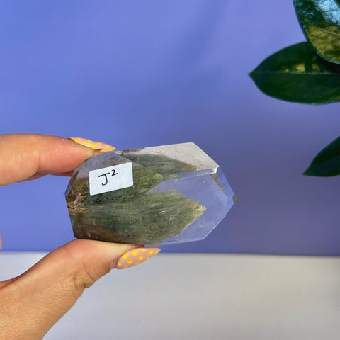 Hand holding a clear, faceted Garden Quartz Free Form with green inclusions and "J2" label.