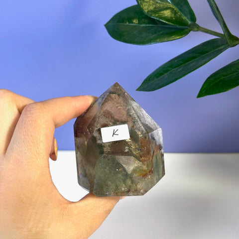 Hand holding a faceted Garden Quartz free form with green and brown inclusions, labeled "K".