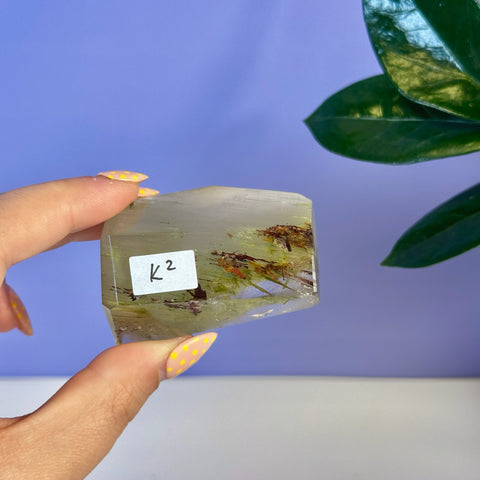 Hand holding a clear Garden Quartz free form with green and reddish-brown inclusions.