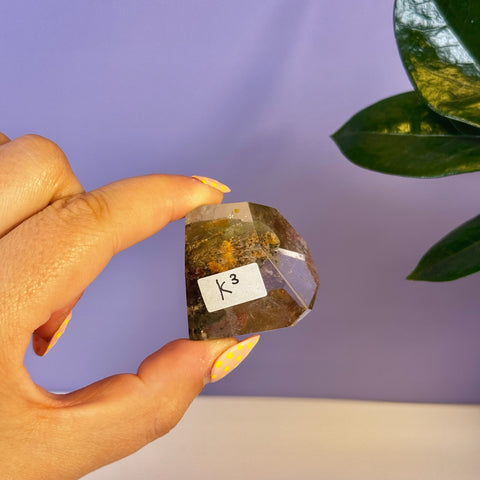 Hand holding a faceted Garden Quartz free form crystal with green and brown inclusions, labeled K3.