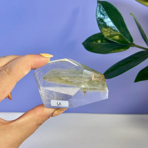 Hand holding a clear Garden Quartz free form with green inclusions and "U" sticker.