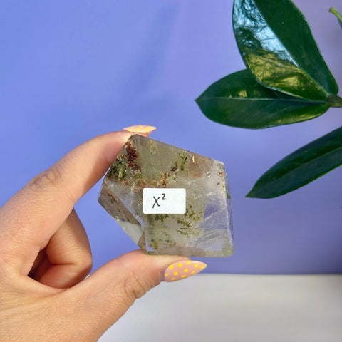 Hand holding a clear Garden Quartz free form crystal with green inclusions and a white X2 label.