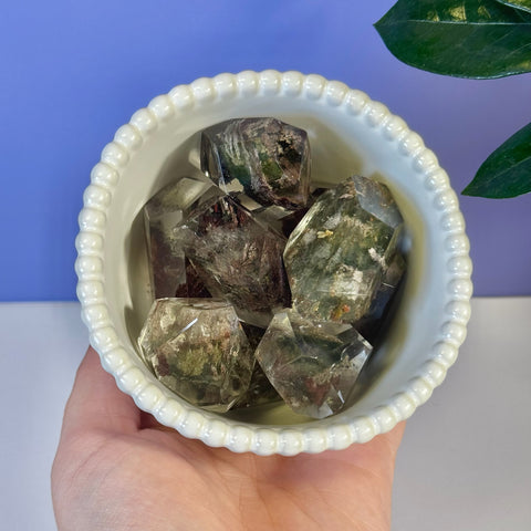 Hand holding a white scalloped bowl filled with several clear and green Garden Quartz Gusher crystals.