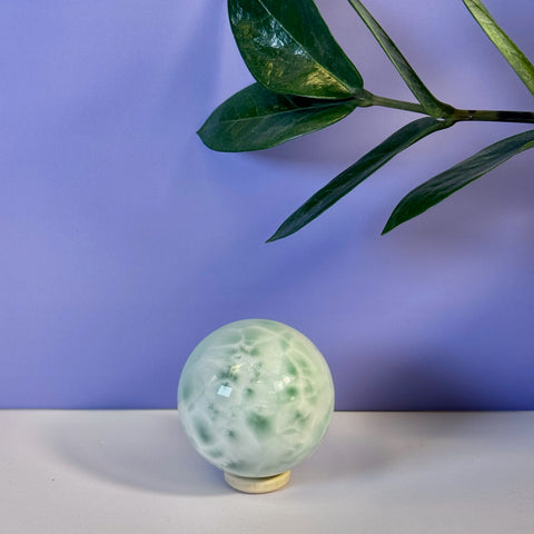 Smooth green alabaster sphere with white marbling on a light wooden stand, set against a purple background with a plant leaf.