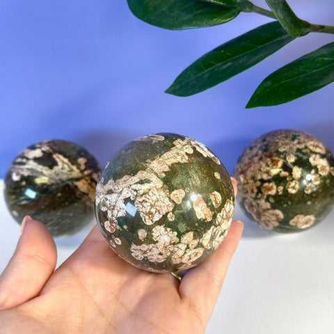 Polished green cherry blossom jasper sphere with white floral patterns, held in a hand.
