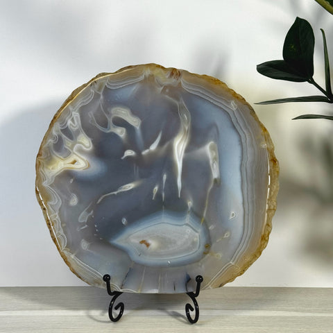 Round, imperfect agate slab with natural grey, white, and brown bands on a black metal stand.