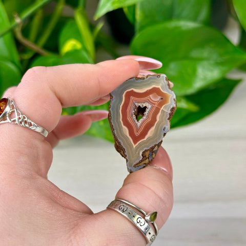 Hand holding a small, polished agate slice with concentric bands of red, orange, white, and gray, featuring a sparkling crystal geode center.