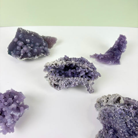 Five clusters of raw, deep purple grape agate with botryoidal, berry-like crystal formations on a white surface.