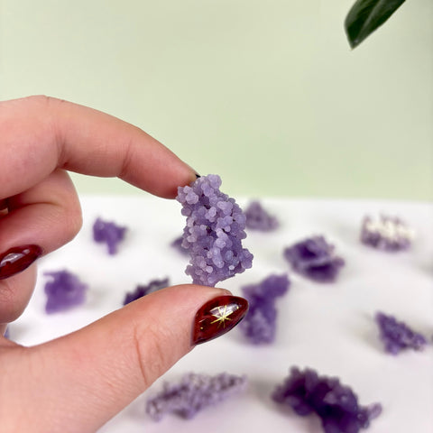 Hand holding a small, bumpy purple grape agate crystal cluster with more pieces scattered on a white surface.