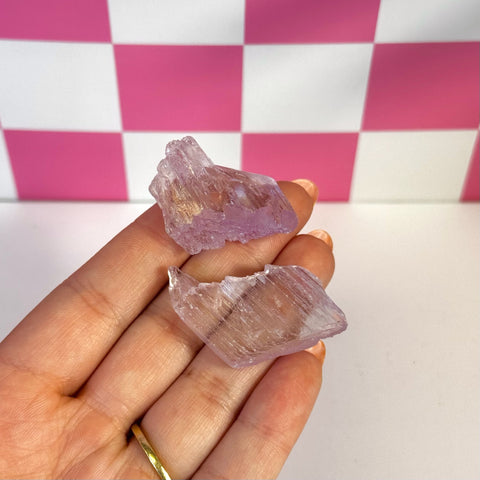 Two raw, pale purple Kunzite crystals held in a hand against a pink and white checkered background.