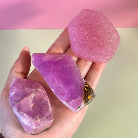 Hand holding three juicy pink aragonite free forms with wave-like patterning and natural inclusions.
