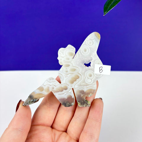 Handheld lounging fairy carving made of translucent scenic moss agate with circular patterns and a small crystal geode.