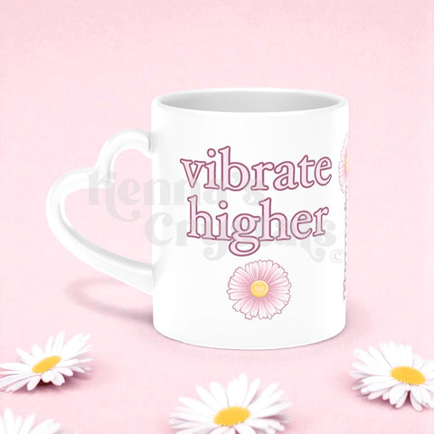 White ceramic mug with heart handle, "vibrate higher" in pink text, and pink daisies on a pink background.