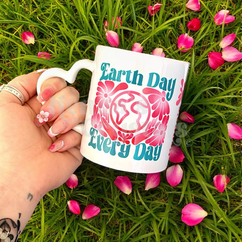 White ceramic mug with "Earth Day Every Day" text, floral design, and heart handle, held in green grass with pink petals.