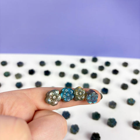 Four small, carved labradorite daisy beads with iridescent blue and gold flashes held on a finger against a purple background.
