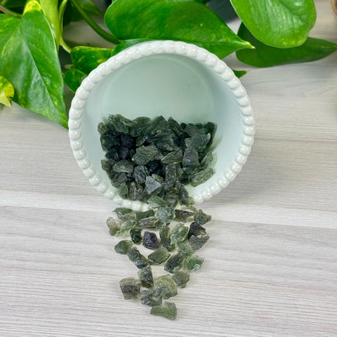 Rough, dark green Moldavite pieces spilling from a white scalloped bowl onto a light wood surface.