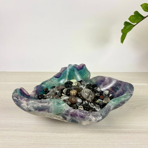 Hand-carved fluorite bowl featuring purple and green hues filled with a variety of polished crystal gemstones.