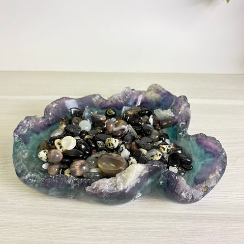 Hand-carved fluorite bowl with natural purple and green hues, filled with a variety of polished dark gemstone crystals.