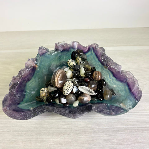 Raw fluorite crystal bowl featuring purple and teal hues, filled with a variety of polished gemstone tumbles.