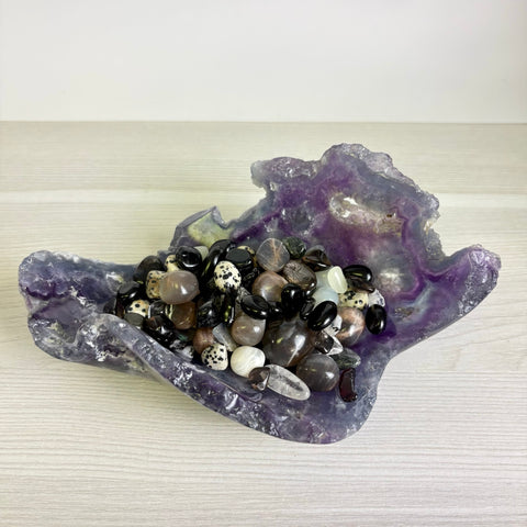 Raw purple fluorite crystal bowl filled with a variety of polished black, white, and spotted gemstone tumbles.