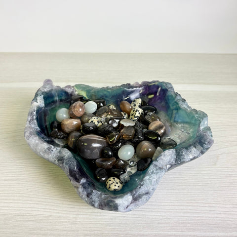 Polished fluorite crystal bowl in shades of purple and green, filled with a collection of assorted smooth gemstone pebbles.