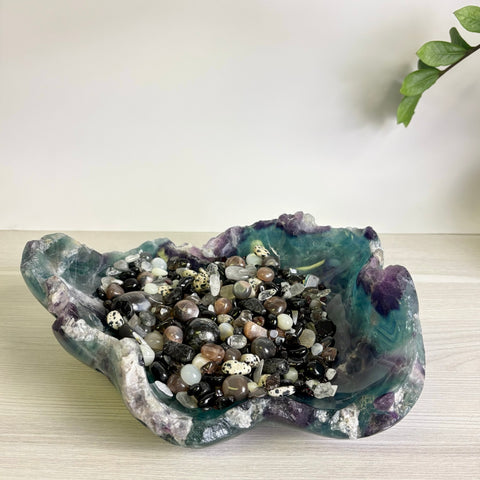 Hand-carved fluorite bowl featuring purple and green hues, filled with a variety of polished black and white crystal stones.