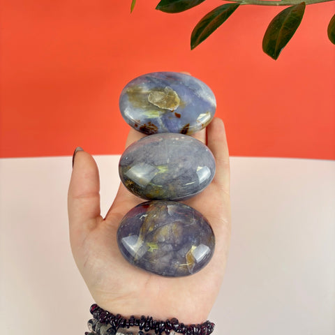 Three smooth, polished Nightshade Chalcedony palm stones with purple and brown mineral inclusions held in a hand.