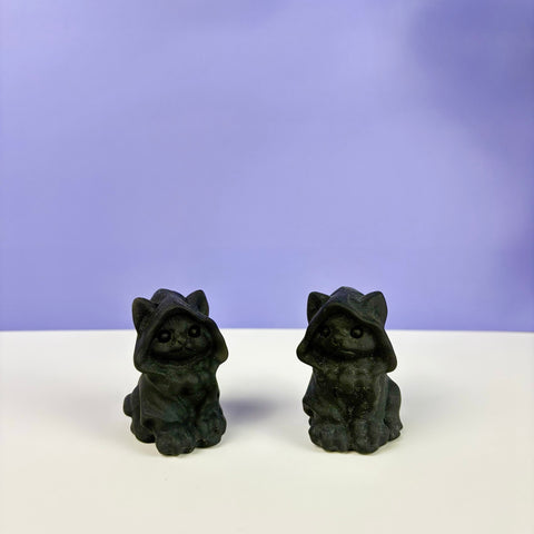 Two small, carved black obsidian cat figurines wearing hooded cloaks on a white surface against a purple background.