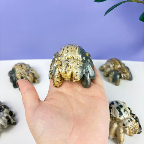 Handheld spider carving made of polished, tan and grey speckled natural stone with detailed legs.