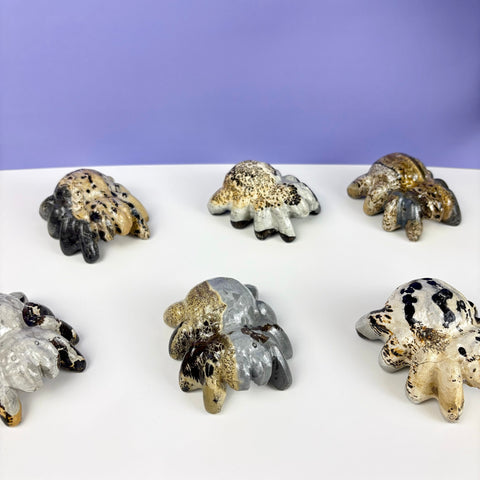 Six hand-painted stone spider figurines in various speckled, metallic, and neutral earth-tone color patterns.