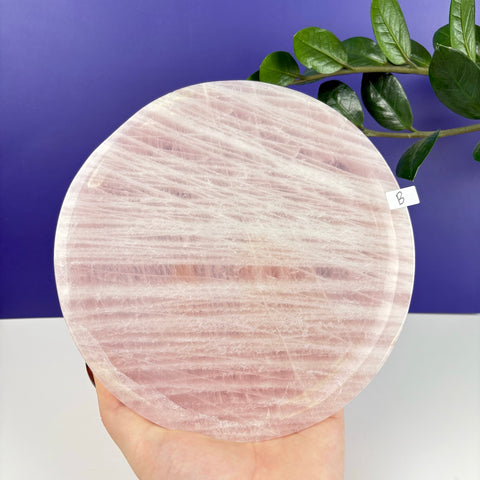 Round rose quartz plate with natural pink veining held in a hand against a purple background.