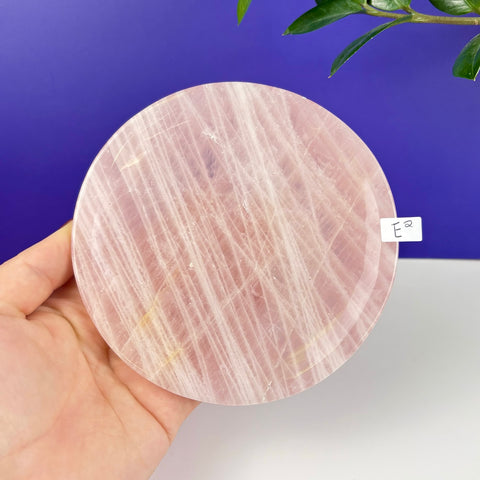 Round rose quartz crystal plate with natural pink veining held in a hand against a blue and white background.