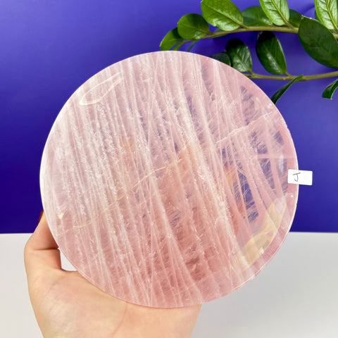Round rose quartz crystal plate with natural pink striations held in a hand against a purple background.