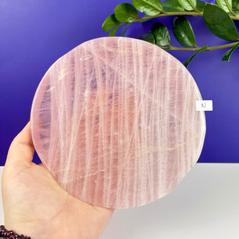 Handheld round rose quartz plate with natural pink crystalline textures and a polished finish against a purple background.