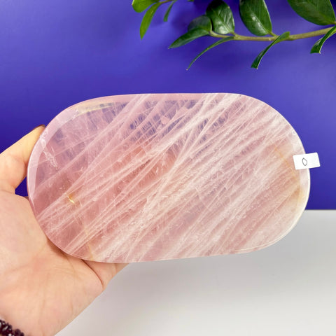 Oval-shaped rose quartz plate with natural pink crystalline textures held in a hand against a blue background.
