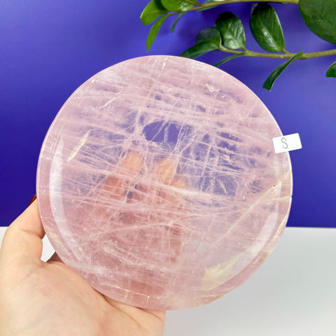 Small round rose quartz crystal plate with natural veining held in a hand against a blue background.