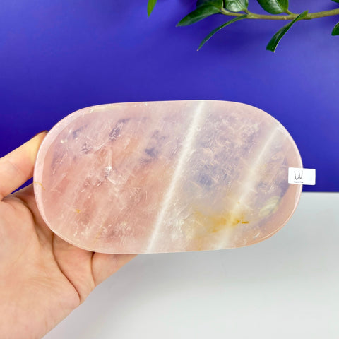 Oval rose quartz crystal tray with natural pink hues held in a hand against a blue and white background.