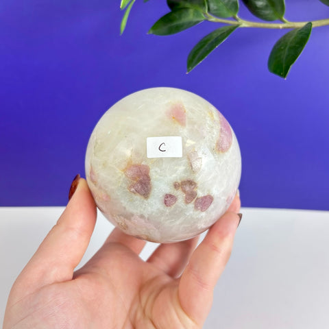 Polished pink tourmaline sphere with natural inclusions held in a hand against a blue and white background.