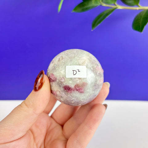 Polished pink tourmaline sphere with green mineral inclusions held in a hand against a blue background.