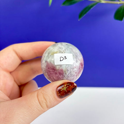 Polished pink tourmaline sphere held in hand against a blue background, featuring natural mineral inclusions and a D3 label.