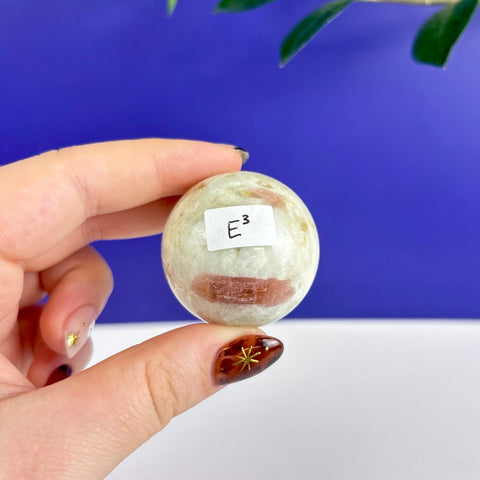 Polished pink tourmaline sphere with white mineral inclusions held in a hand against a blue and white background.