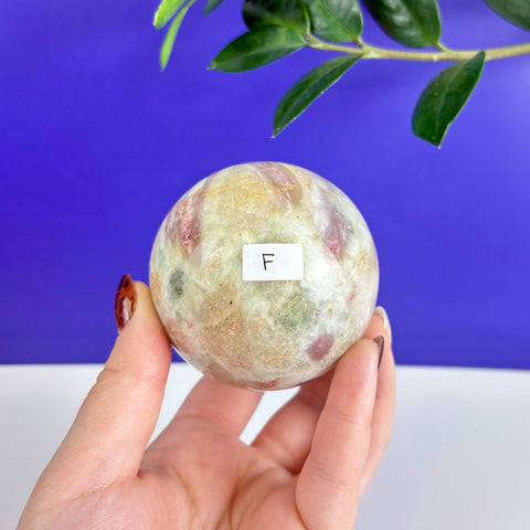 Polished pink tourmaline sphere with natural mineral inclusions held in a hand against a blue and white background.