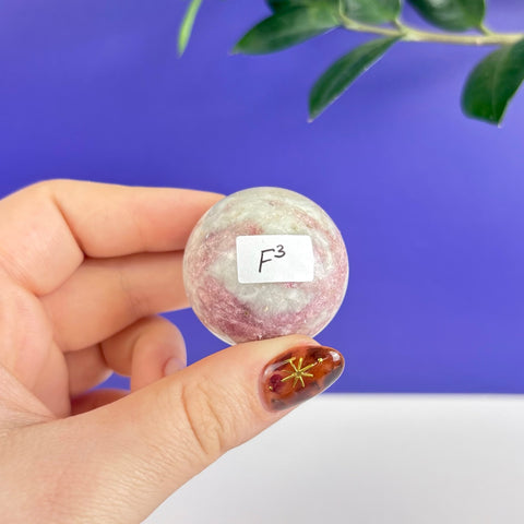 Small polished pink and white tourmaline sphere held in hand against a blue background.