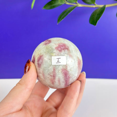 Polished pink tourmaline sphere with white quartz inclusions held in a hand against a blue background.