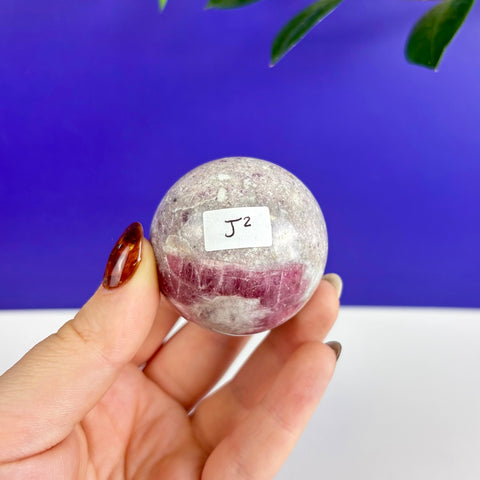 Hand holding a polished pink tourmaline sphere with natural white and magenta mineral inclusions against a blue background.